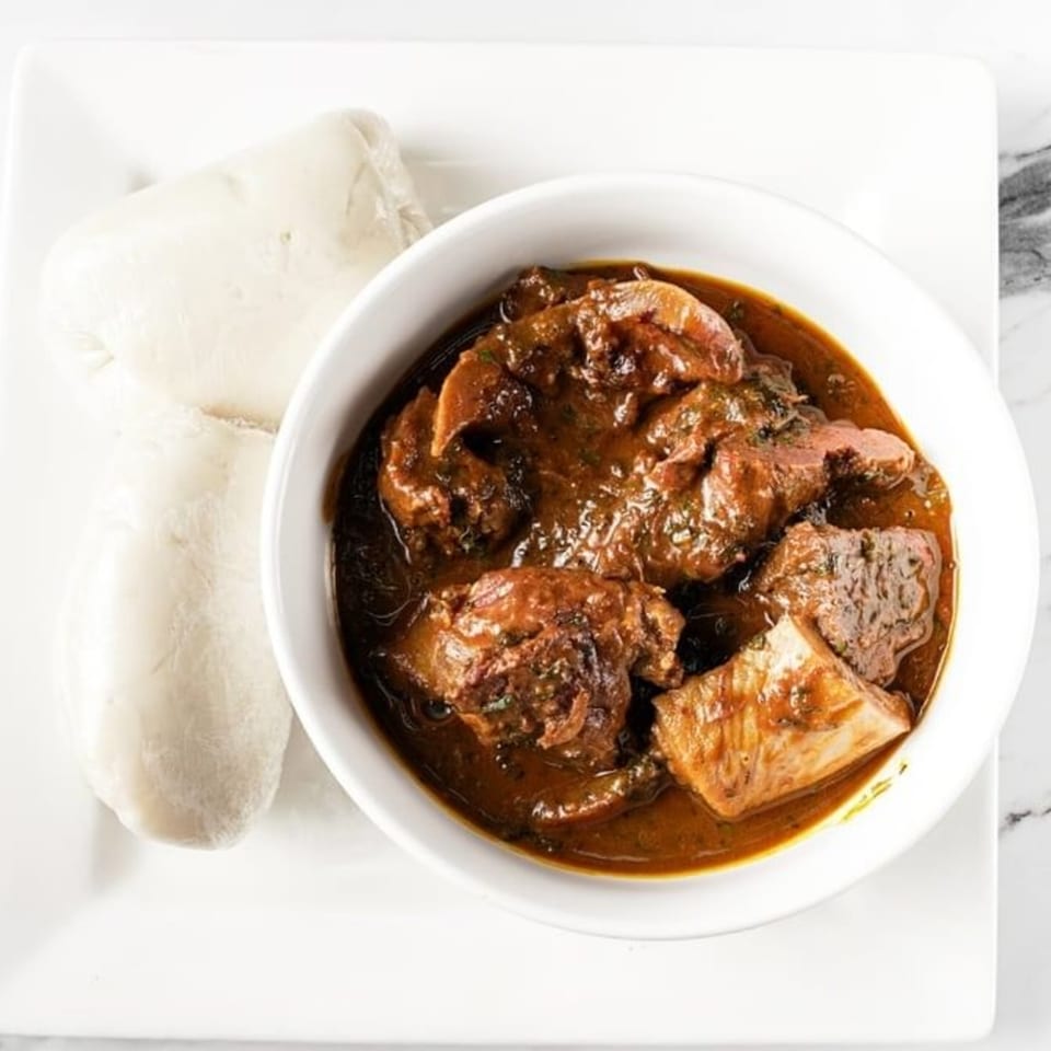 A bowl of dark brown African stew with meat and vegetables, served with two white fufu portions.