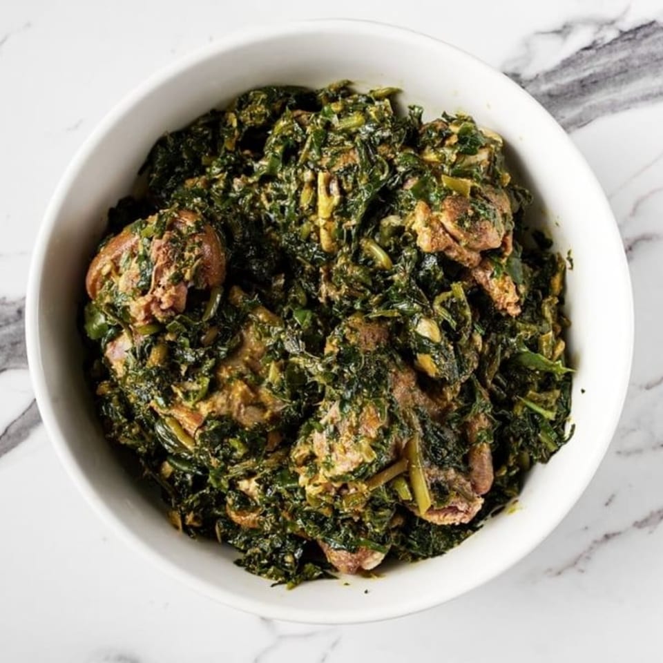 A white bowl filled with a dark green, leafy African stew with pieces of meat.